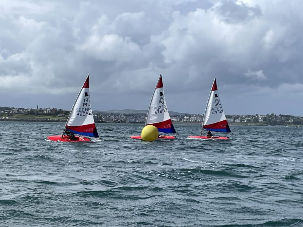 Three Topper dinghies in line sailing right to left and passing a yellow marker