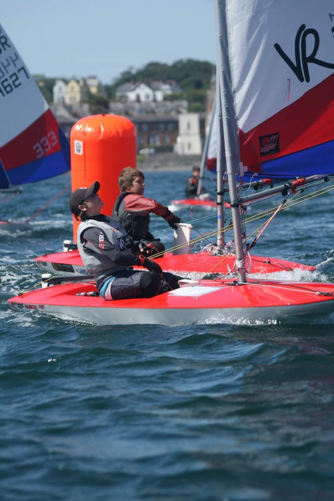 two red topper dinghies with white blue and red sails close to the camera, each sailed by a boy of around 13 years old, round a large orange marker in sea with the town of Donaghadee in the back ground